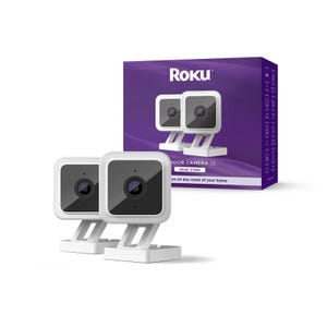2-pack of Roku Indoor Cameras SE in white with a wired setup, featuring a sleek square design and a lens in the center, displayed alongside a matching purple box with product information.