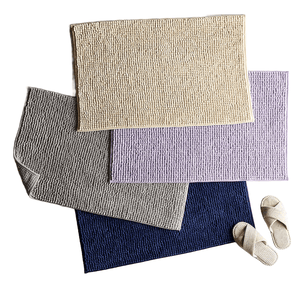 Four textured bath mats in beige, gray, lavender, and dark blue are layered together, accompanied by a pair of light-colored slippers.