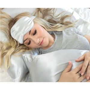 A woman is wearing a smooth, silky, silver Mulberry Silk Eye Mask, resting on a matching silk pillowcase. The mask and pillowcase are crafted from a lustrous fabric designed to enhance comfort during sleep.