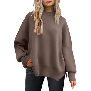 A woman models an oversized brown ribbed sweater paired with dark jeans. The sweater features a high neckline and dropped shoulders.