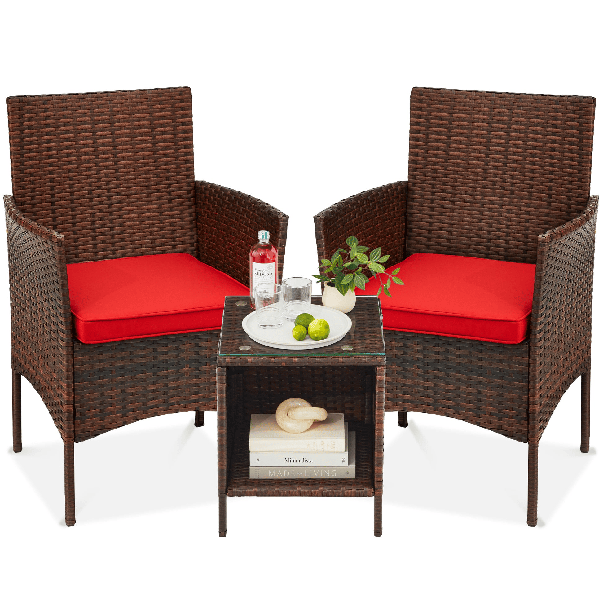 Rattan patio set with two chairs featuring red cushions, and a small glass-top table. The table holds limes, a bottle, and a small plant, with books and a decorative object on the shelf below.