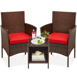 Rattan patio set with two chairs featuring red cushions, and a small glass-top table. The table holds limes, a bottle, and a small plant, with books and a decorative object on the shelf below.