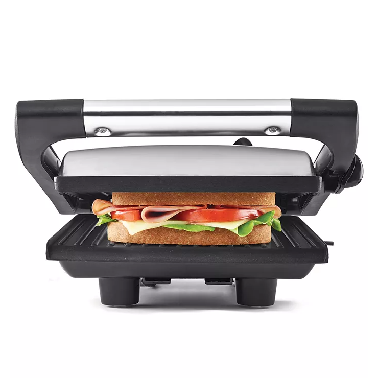 A panini press and sandwich grill featuring a stainless steel top, black handle, and grill plates, shown pressing a sandwich with lettuce, tomato, cheese, and meat.