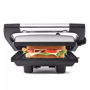 A panini press and sandwich grill featuring a stainless steel top, black handle, and grill plates, shown pressing a sandwich with lettuce, tomato, cheese, and meat.