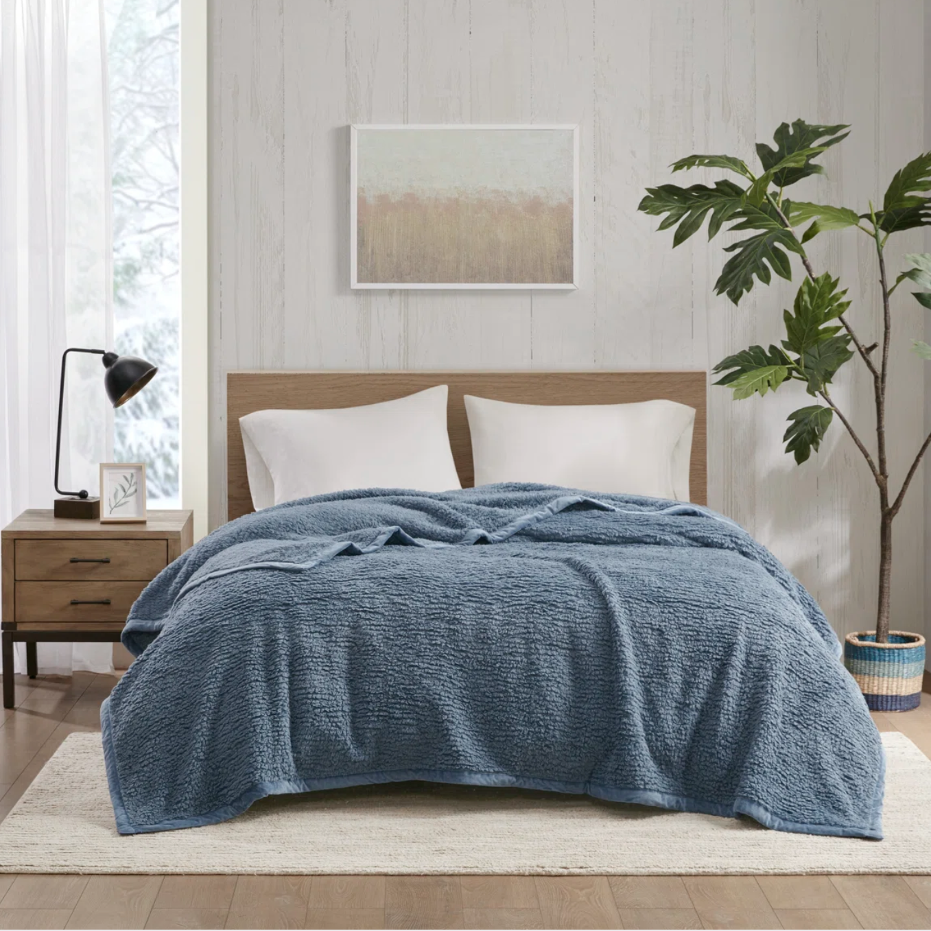 A bedroom features a bed with a blue textured comforter and white pillows, a wooden headboard, a wooden nightstand with a black lamp, a framed wall art piece, and a potted leafy plant on a rug, all in a minimalist style.