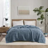 A bedroom features a bed with a blue textured comforter and white pillows, a wooden headboard, a wooden nightstand with a black lamp, a framed wall art piece, and a potted leafy plant on a rug, all in a minimalist style.