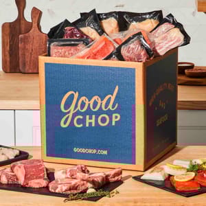 Good Chop offers a delivery box containing various vacuum-sealed meats, including steaks and seafood, displayed on a wooden kitchen counter.
