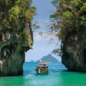 A wooden boat travels between two rocky cliffs covered with greenery in turquoise waters, with a distant island visible under a clear blue sky.