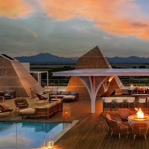 A stylish rooftop setting with woven pyramid structures, lounge seating, a bar with stools, a fire pit, and a pool, all under a colorful sunset sky.