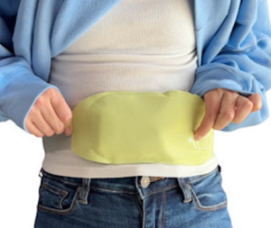 A person wears a light green heating pad wrapped around their lower abdomen, secured under a blue jacket and over a white shirt.