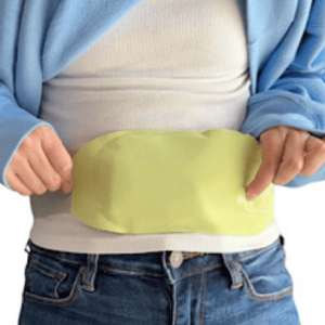 A person wears a light green heating pad wrapped around their lower abdomen, secured under a blue jacket and over a white shirt.