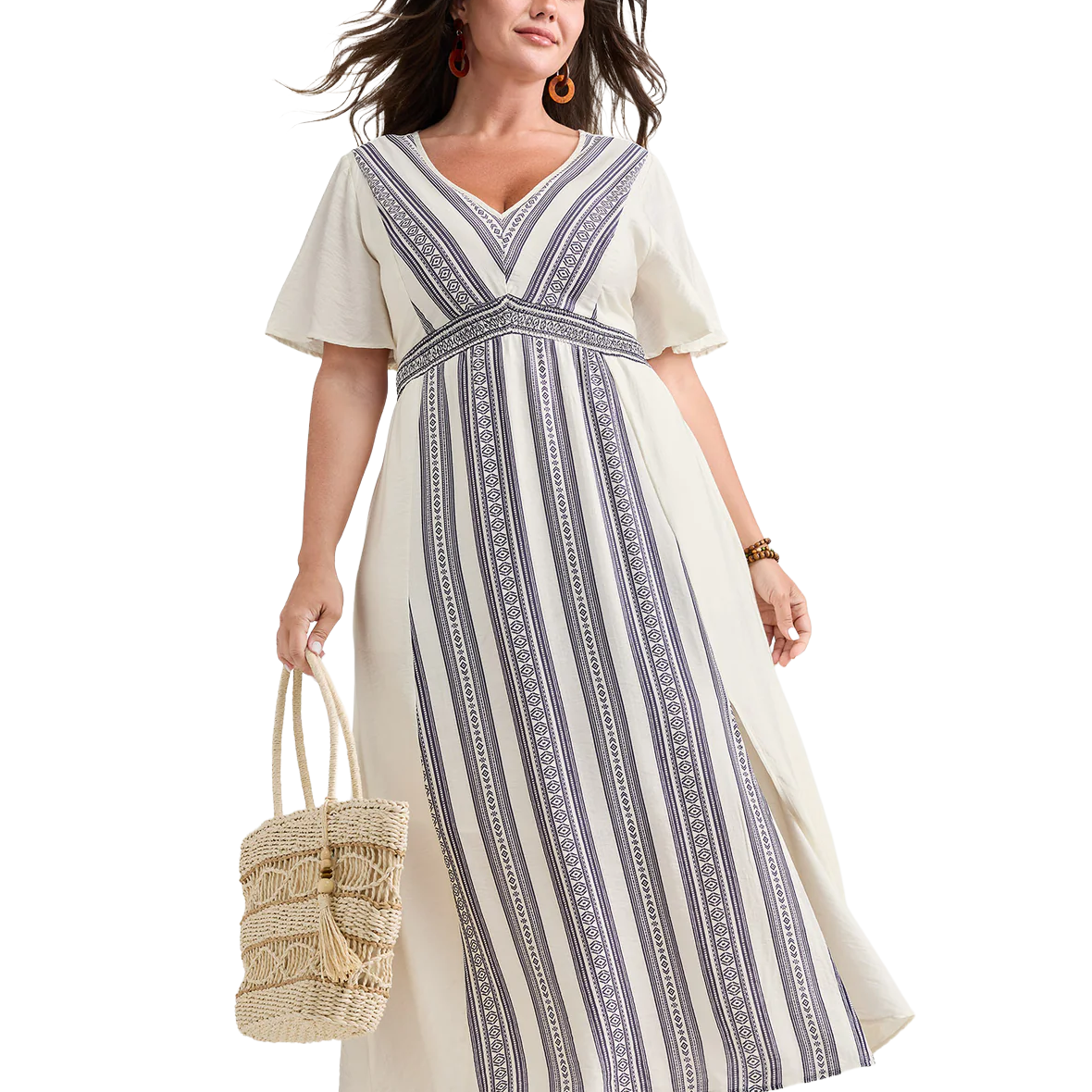 A woman wears a white maxi dress featuring navy geometric vertical stripes and short sleeves, accessorized with a woven straw bag and orange earrings.