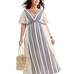A woman wears a white maxi dress featuring navy geometric vertical stripes and short sleeves, accessorized with a woven straw bag and orange earrings.