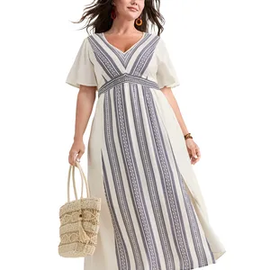 A woman wears a white maxi dress featuring navy geometric vertical stripes and short sleeves, accessorized with a woven straw bag and orange earrings.