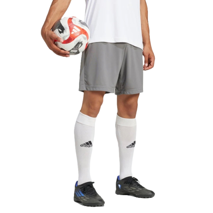 A person wearing gray athletic shorts, a white shirt, and white knee-high socks with the Adidas logo, holding a soccer ball. They are also wearing black Adidas soccer cleats with blue accents.