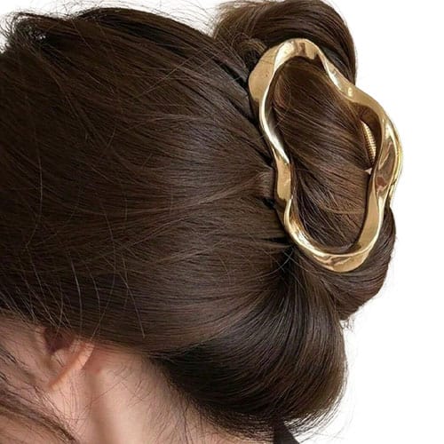 A person with brown hair styled in a bun is securing it with a gold hair clip featuring an irregular, wavy design.