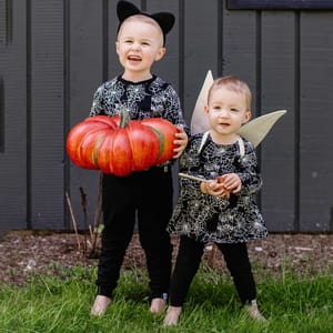 Two children in Halloween-themed outfits, including cat ears and spider-web patterns, hold a large decorative pumpkin. One child has wings and holds a wand.