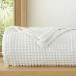 A folded white waffle-patterned blanket on a wooden surface, with a large window and greenery visible in the background.
