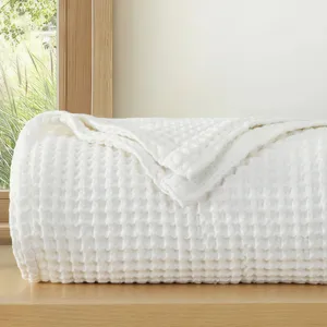 A folded white waffle-patterned blanket on a wooden surface, with a large window and greenery visible in the background.
