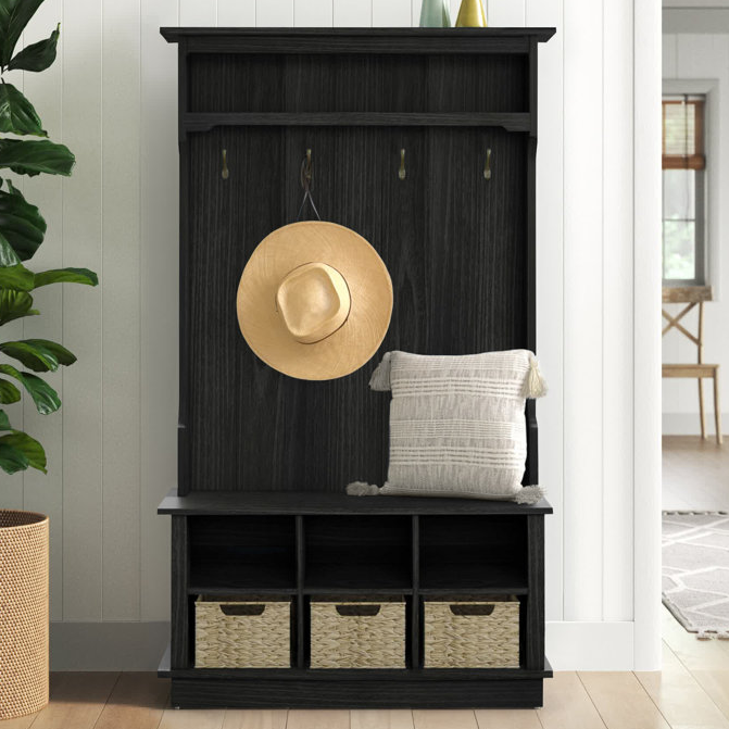 A black hall tree with a built-in bench, featuring three hooks for hanging items and three cubbies with woven baskets for storage. A pillow and a hat are displayed on the bench.
