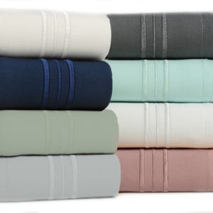 A stack of rolled bamboo sheet sets in six colors: cream, gray, navy, light turquoise, sage, and rose, each featuring decorative triple-stitched bands.