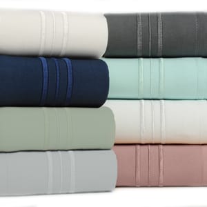 A stack of rolled bamboo sheet sets in six colors: cream, gray, navy, light turquoise, sage, and rose, each featuring decorative triple-stitched bands.