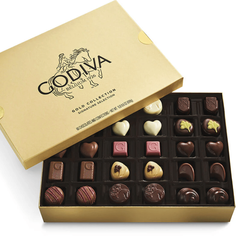 A gold box labeled \“Godiva Belgium 1926 Gold Collection Signature Selection\“ contains assorted chocolates with various shapes like hearts and squares.
