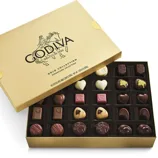 A gold box labeled \“Godiva Belgium 1926 Gold Collection Signature Selection\“ contains assorted chocolates with various shapes like hearts and squares.