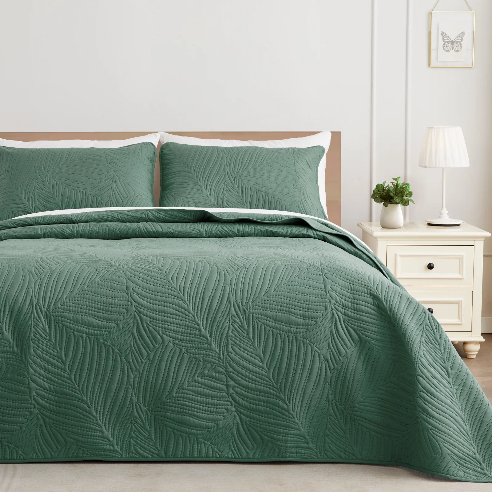 A green quilted bed cover set with leaf patterns, including two matching pillowcases, on a bed next to a nightstand with a lamp and plant.