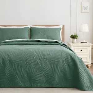 A green quilted bed cover set with leaf patterns, including two matching pillowcases, on a bed next to a nightstand with a lamp and plant.