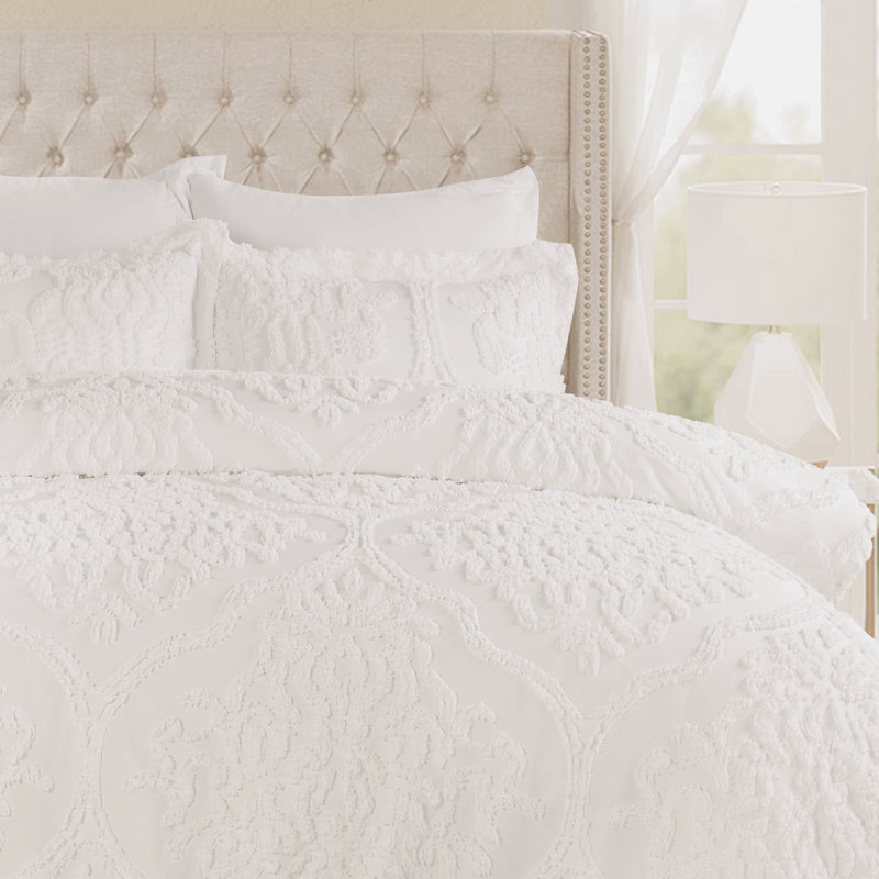 A white 7-piece queen comforter set featuring a boho textured design includes a comforter, two shams, two decorative pillows, and two euro shams. The bedding is displayed on a plush, tufted headboard with a side lamp and window curtain in the background.