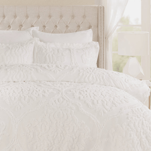 A white 7-piece queen comforter set featuring a boho textured design includes a comforter, two shams, two decorative pillows, and two euro shams. The bedding is displayed on a plush, tufted headboard with a side lamp and window curtain in the background.