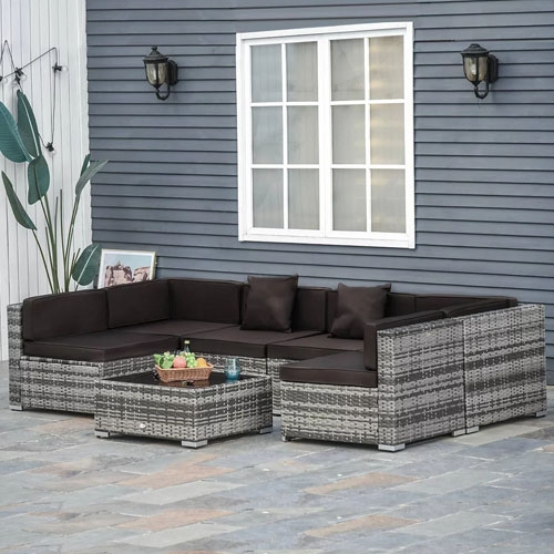 Outdoor patio set featuring a sectional sofa and coffee table in gray rattan with dark cushions, placed on a stone tile floor against a blue-gray wall with windows and decorative lamps.