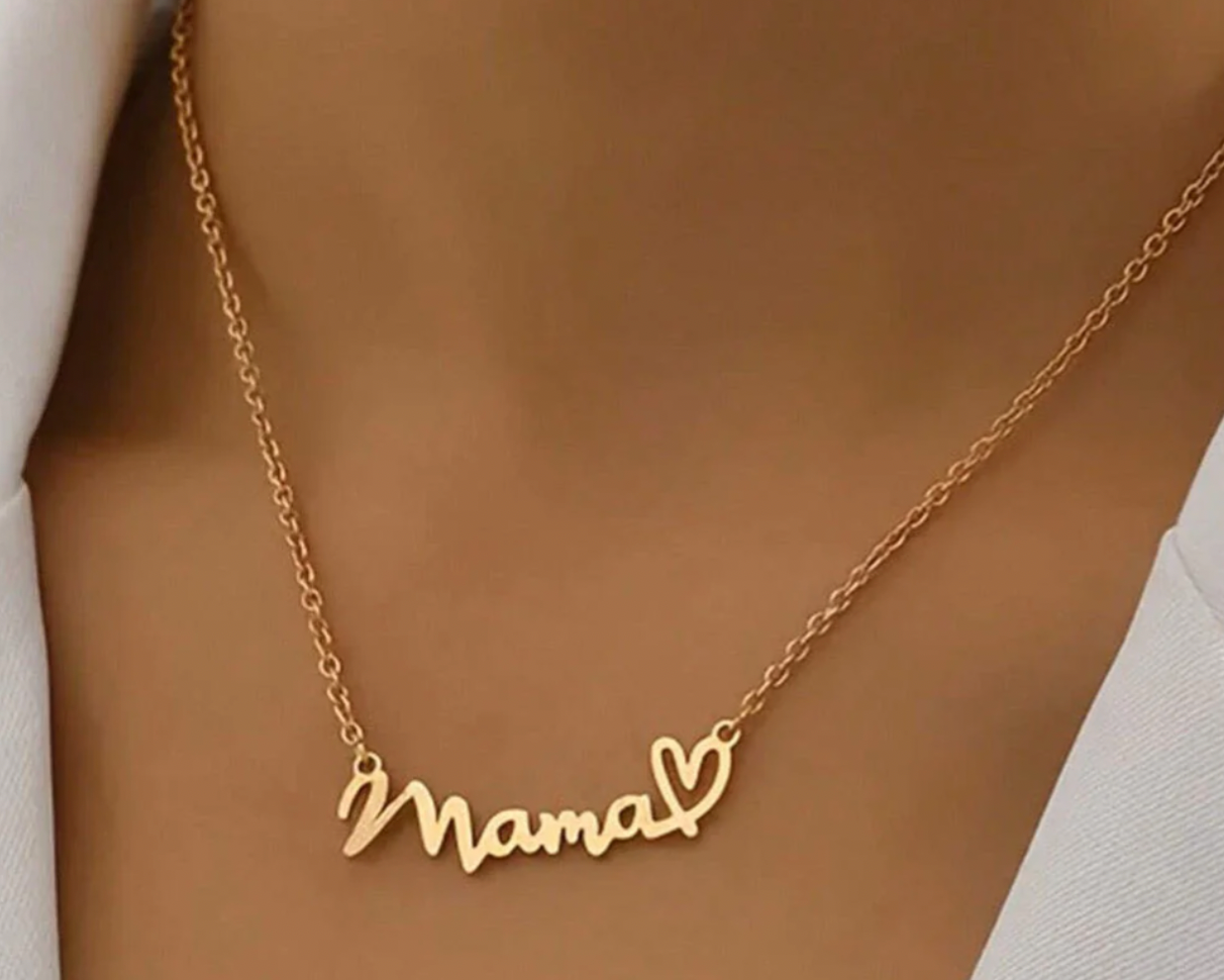 A gold necklace featuring the word \“Mama\“ in cursive script, adorned with a heart symbol at the end, hanging on a delicate chain around the neck.