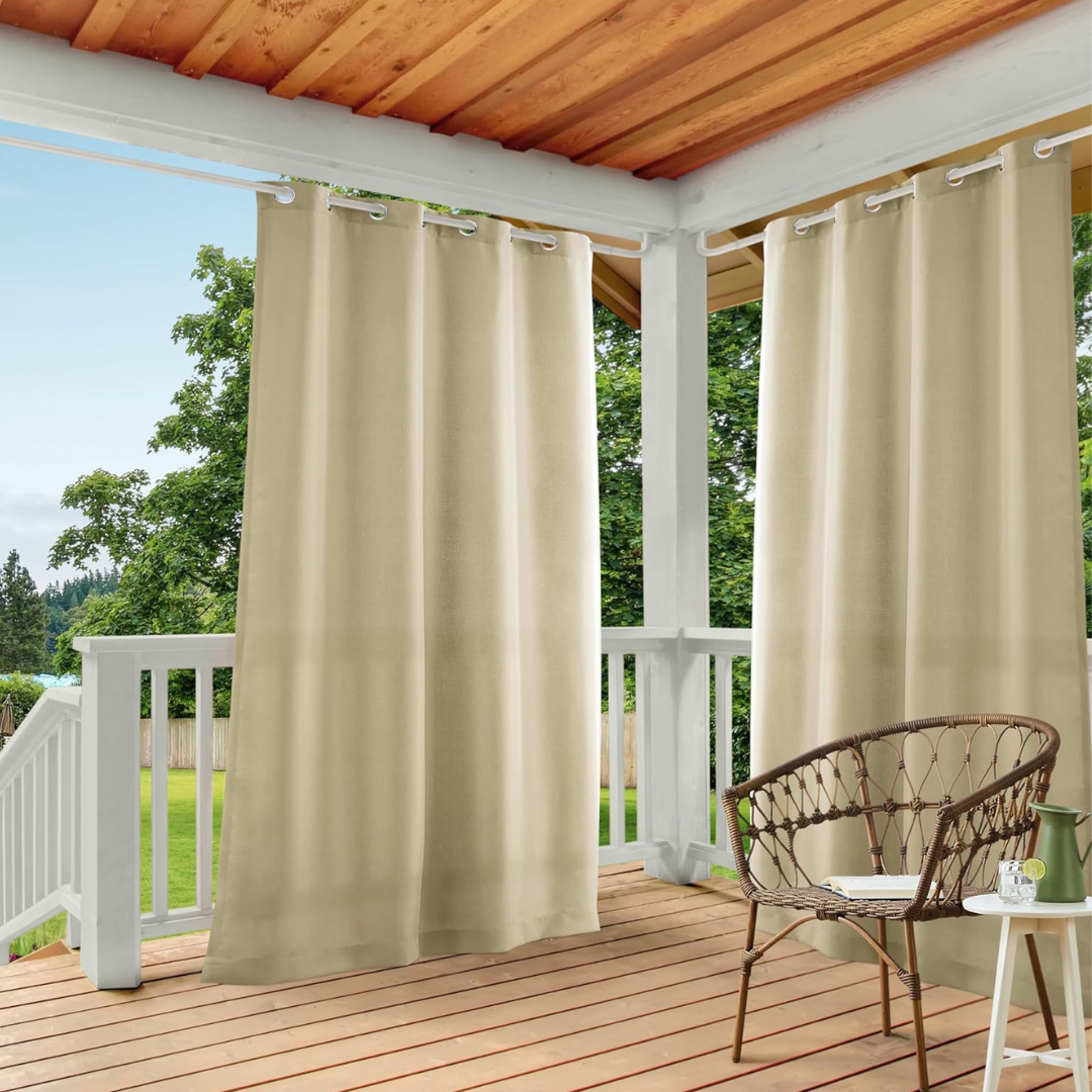 Outdoor beige curtains with grommets hang on a porch, adjacent to a wicker chair and a small table with a book and glasses, against a backdrop of greenery.