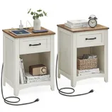 Two white nightstands with wooden tops, featuring a drawer, open storage space, and a built-in charging station. One holds a vase, tablet, and books, while the other has an alarm clock and basket. Both are connected with visible power cords.
