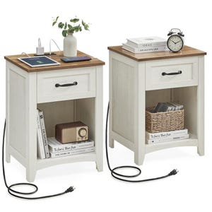 Two white nightstands with wooden tops, featuring a drawer, open storage space, and a built-in charging station. One holds a vase, tablet, and books, while the other has an alarm clock and basket. Both are connected with visible power cords.