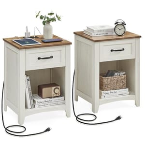 Two white nightstands with wooden tops, featuring a drawer, open storage space, and a built-in charging station. One holds a vase, tablet, and books, while the other has an alarm clock and basket. Both are connected with visible power cords.