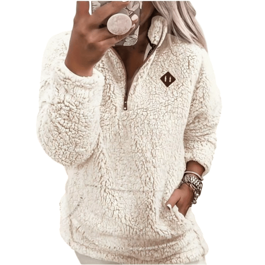 A person wears a cream-colored, plush, sherpa pullover with a quarter-zip front and a small emblem. The outfit includes a matching phone case, watch, and bracelets.