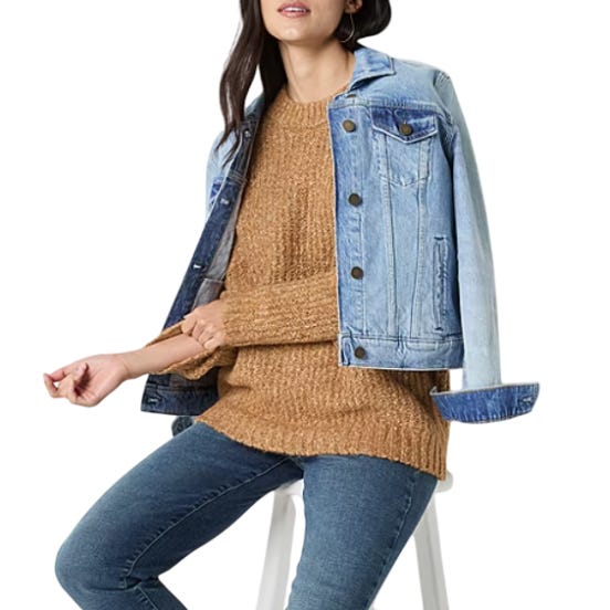 A woman is wearing a brown knit sweater, blue denim jeans, and a light blue denim jacket draped over her shoulders, while seated on a stool.