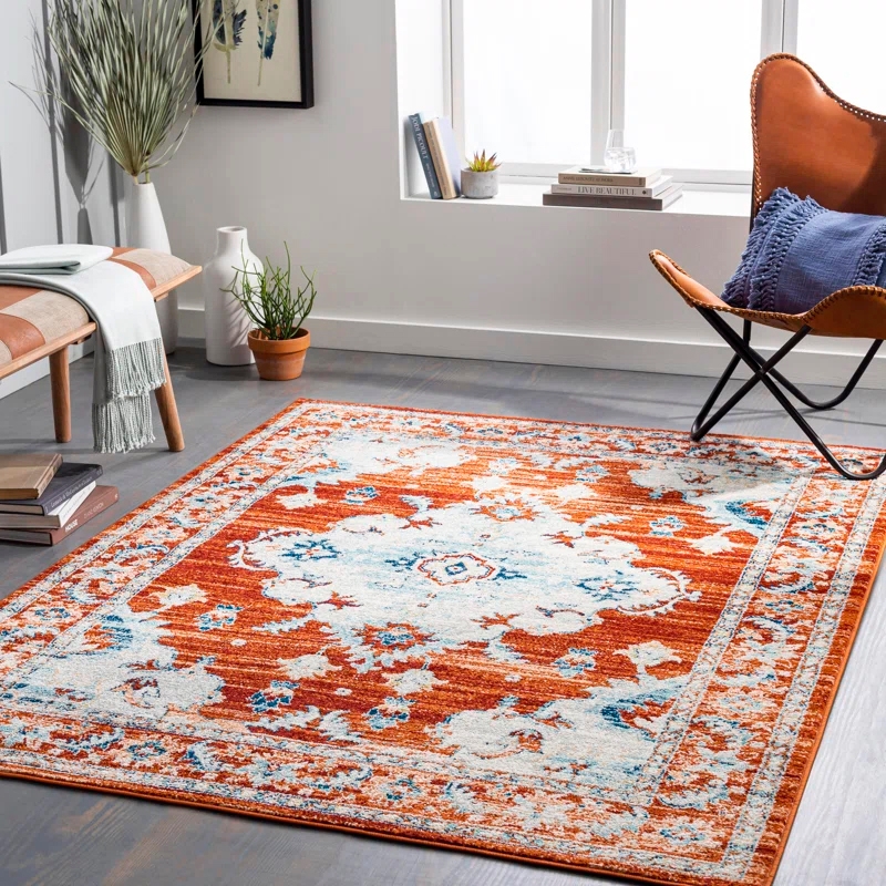The 7' x 9' Aldusa Area Rug features an intricate floral pattern with orange, blue, and white hues. It's placed in a well-lit room with modern furniture, including a leather chair and a wooden bench.