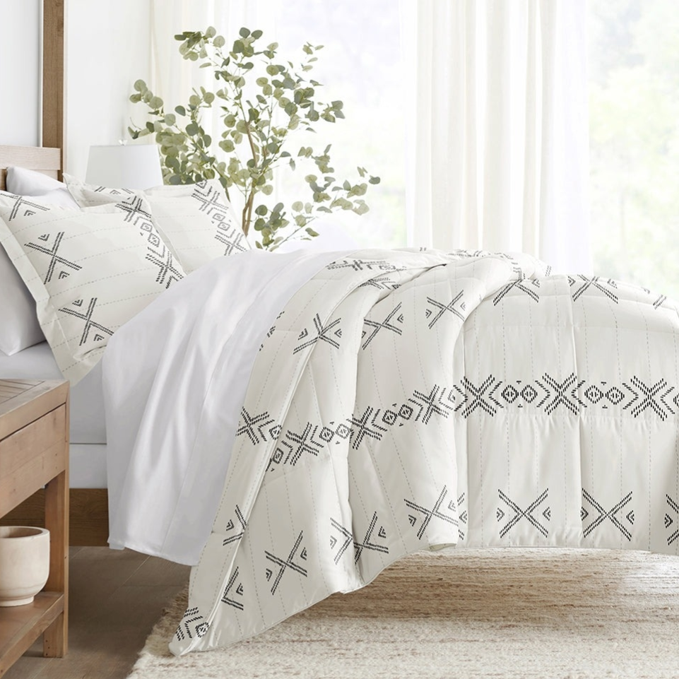 A bed is adorned with a white comforter set featuring a black geometric pattern, accompanied by matching pillow shams. A wooden side table and a potted plant are nearby.