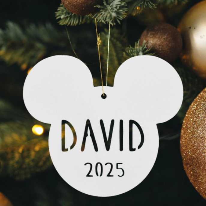 A Mickey Mouse-shaped ornament with the name \“David\“ and the year \“2025\“ hangs on a Christmas tree amid gold and bronze baubles.