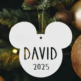 A Mickey Mouse-shaped ornament with the name \“David\“ and the year \“2025\“ hangs on a Christmas tree amid gold and bronze baubles.