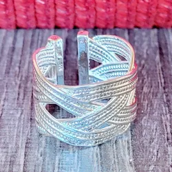 A silver ring with an intricate criss-cross design, featuring textured detailing and an open back, is displayed on a wooden surface with a red background.