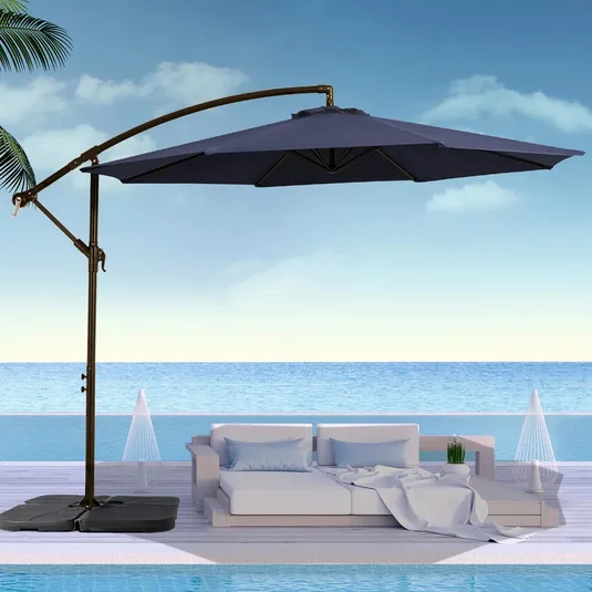 A blue cantilever patio umbrella provides shade over a modern outdoor sofa set by the pool, with a background of ocean and sky.