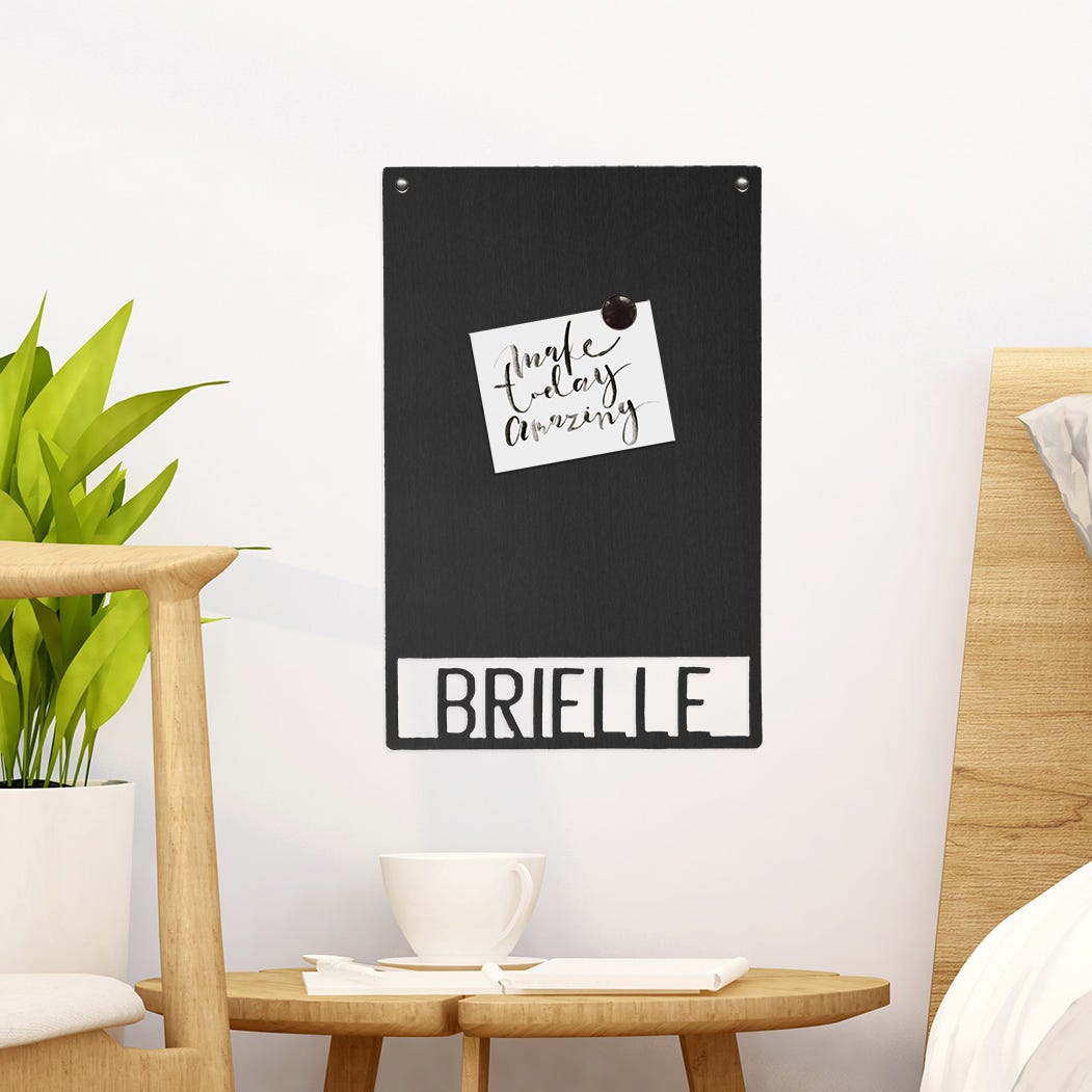 A black chalkboard wall organizer with the name \“Brielle\“ at the bottom, featuring a note pinned on it saying \“make today amazing.\“ The setting includes a plant, a wooden chair, and a cup on a small round table.