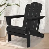 A black Adirondack chair is placed on a light wood floor next to potted plants and a white vase, set against a textured white wall.