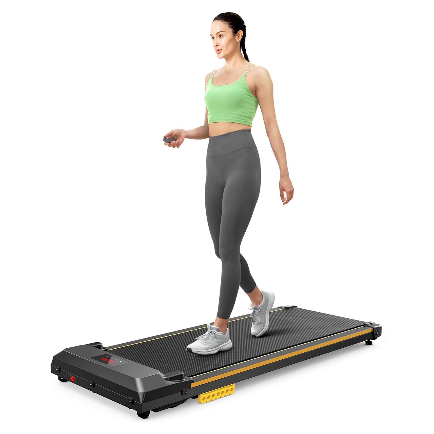 A woman walks on a compact, black walking pad treadmill with a digital display, wearing gray leggings and a green crop top, holding a remote control. The treadmill features a slim design suitable for home use.