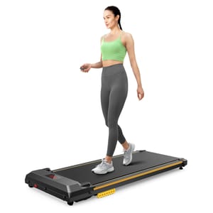 A woman walks on a compact, black walking pad treadmill with a digital display, wearing gray leggings and a green crop top, holding a remote control. The treadmill features a slim design suitable for home use.